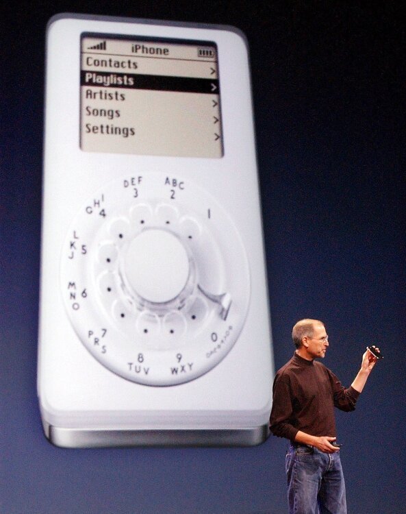 Steve Jobs stands in front of a spoof image of an iPod with a rotary dial, pretending it