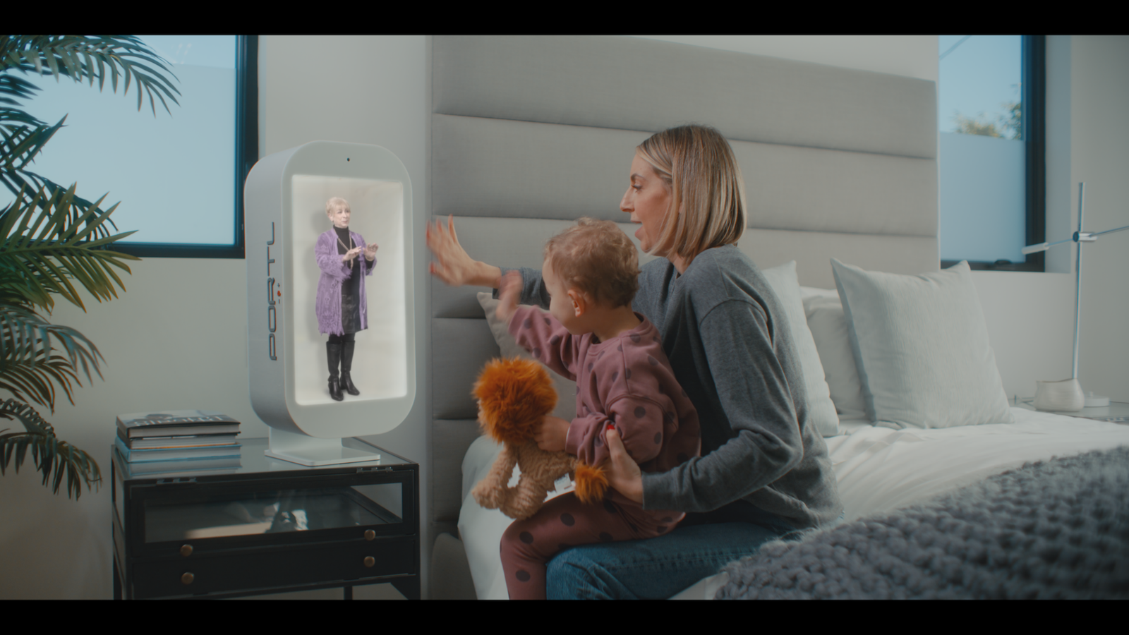 A woman with a kid on her lap sitting on a bed waving to a screen on the bedside table.